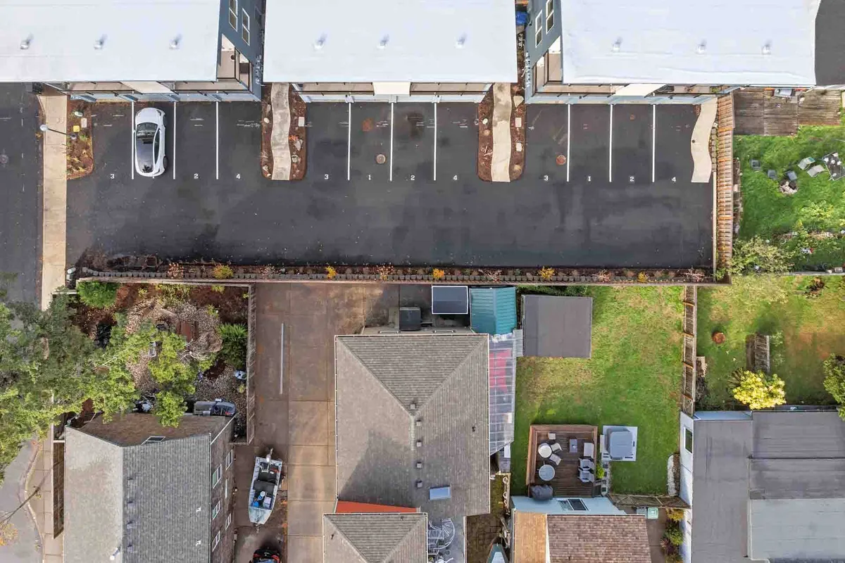 An exterior view featuring neatly arranged parking spaces, a green backyard, and the rooftops of neighboring houses. The scene has a community-oriented and functional mood, suggesting easy access to outdoor areas and the surrounding neighborhood.