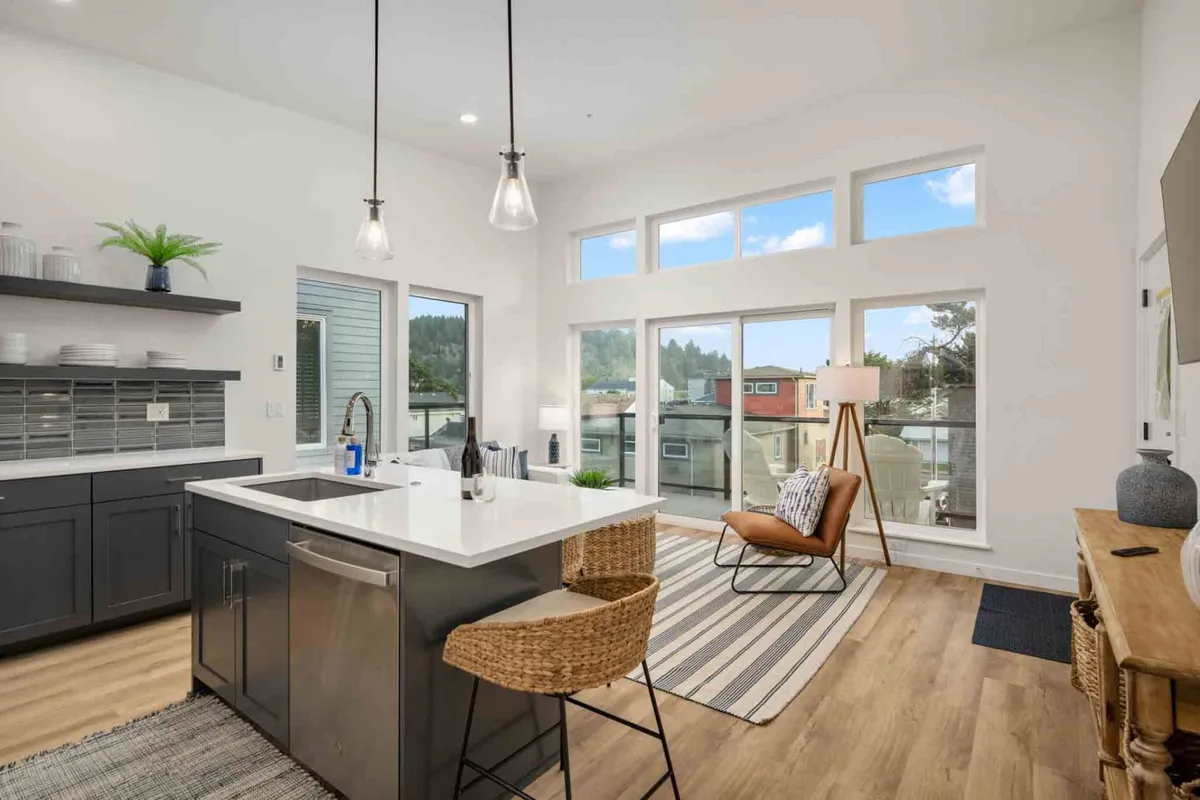 A bright interior space featuring a kitchen island, large windows that flood the room with natural light, and a wooden chair placed in an inviting corner. The sunlight enhances the clean lines and modern coastal aesthetic, with subtle wood tones complementing the simple yet elegant decor.