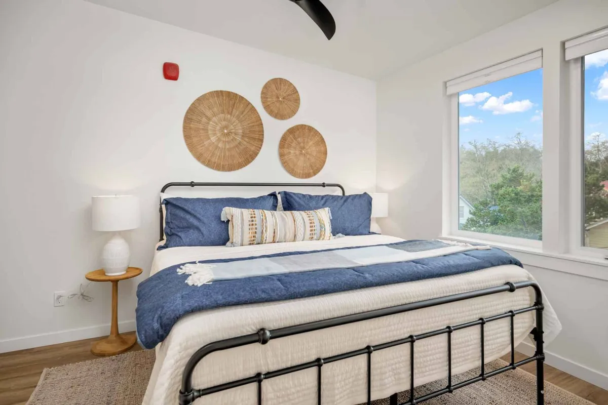 A tranquil bedroom with a comfortable bed adorned with soft linens, framed by tasteful wall decor and illuminated by natural light from the windows. A nightstand and lamp add functional elegance, creating an inviting space for rest and relaxation.