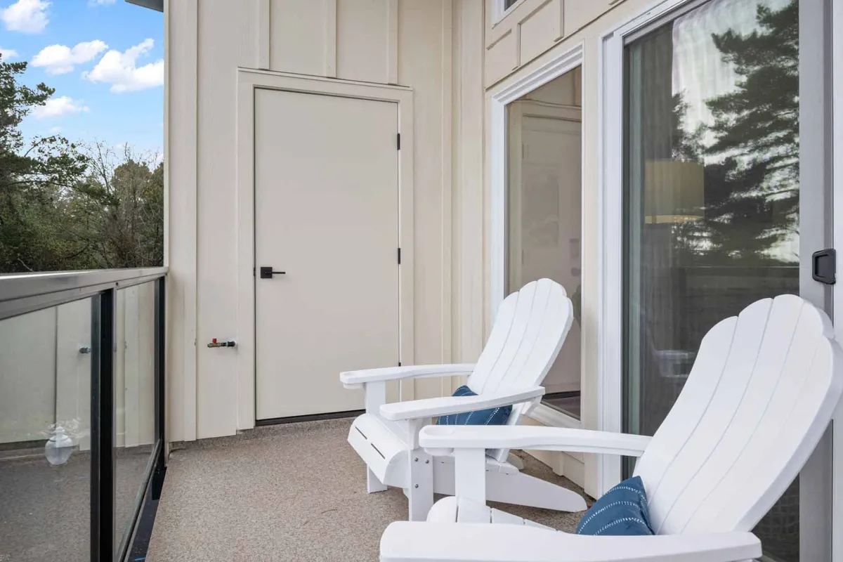 This image features two white Adirondack chairs positioned on a balcony with a glass railing, a door leading inside, and an inviting, tranquil exterior setting. The scene is bathed in soft, natural light, suggesting a relaxed atmosphere ideal for morning coffee, reading, or unwinding at sunset.
