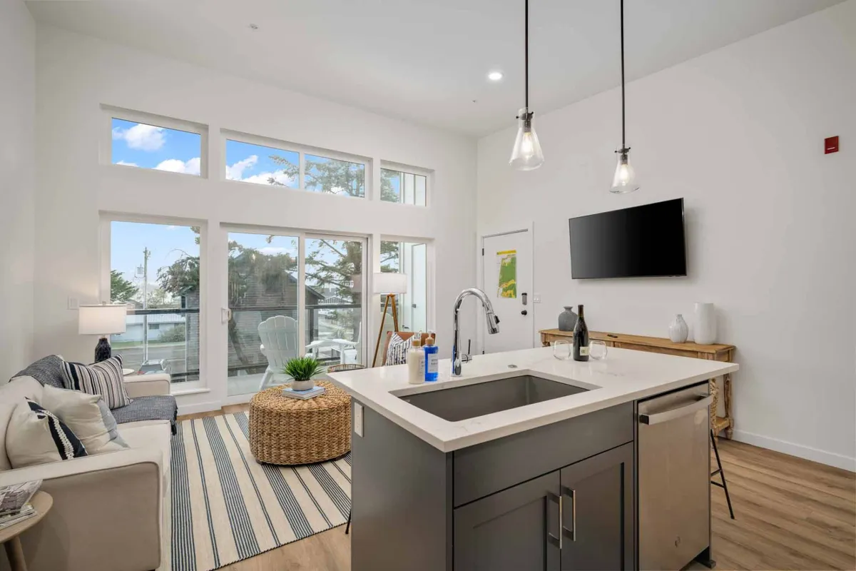 The image showcases the heart of the condo—a stylish, sun-filled open-plan living space featuring a modern kitchen island, plush seating, and expansive windows that flood the room with natural light. The large windows offer tranquil views and invite the colors of the Oregon coast inside, while the wall-mounted TV and inviting sofa create a space perfect for gathering, relaxing, or entertaining.