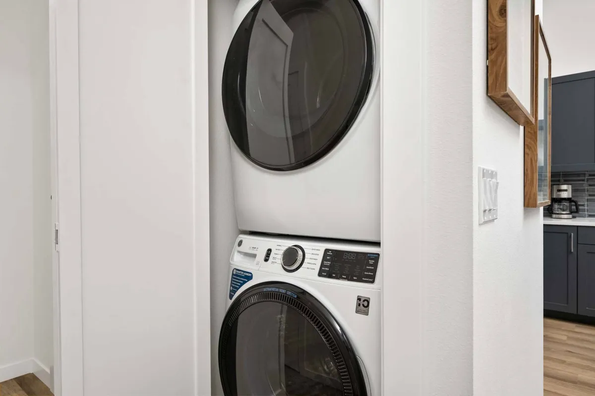 A practical interior scene showing a stacked washer and dryer, a framed mirror, and part of the kitchen in the background. The space is modern and efficient, supporting day-to-day convenience for long-term or remote stays.