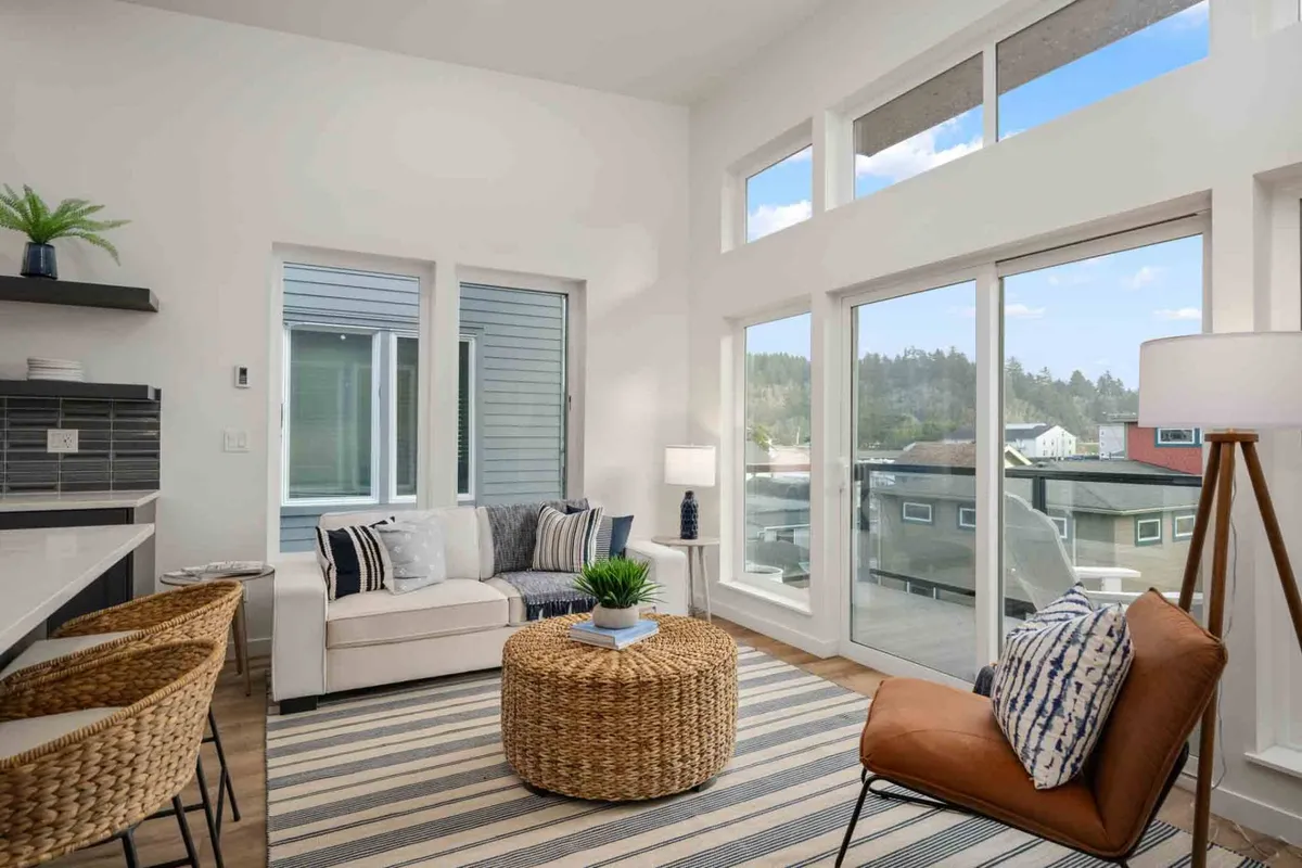 A cozy interior lounge area with large windows, a textured area rug, a woven coffee table, and an inviting armchair. The space is filled with light, offering a serene spot to unwind or entertain, with views reaching out towards the neighborhood below.