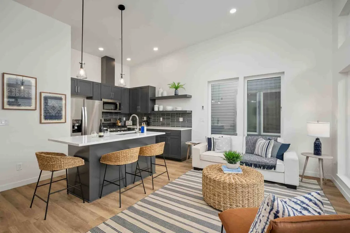 This image features a welcoming open-concept space that seamlessly combines a modern kitchen with a central island, a plush sofa for lounging, decorative plants that add warmth, and bar stools inviting casual conversation. Ample natural light flows through the area, highlighting the inviting atmosphere ideal for both relaxation and social gatherings.