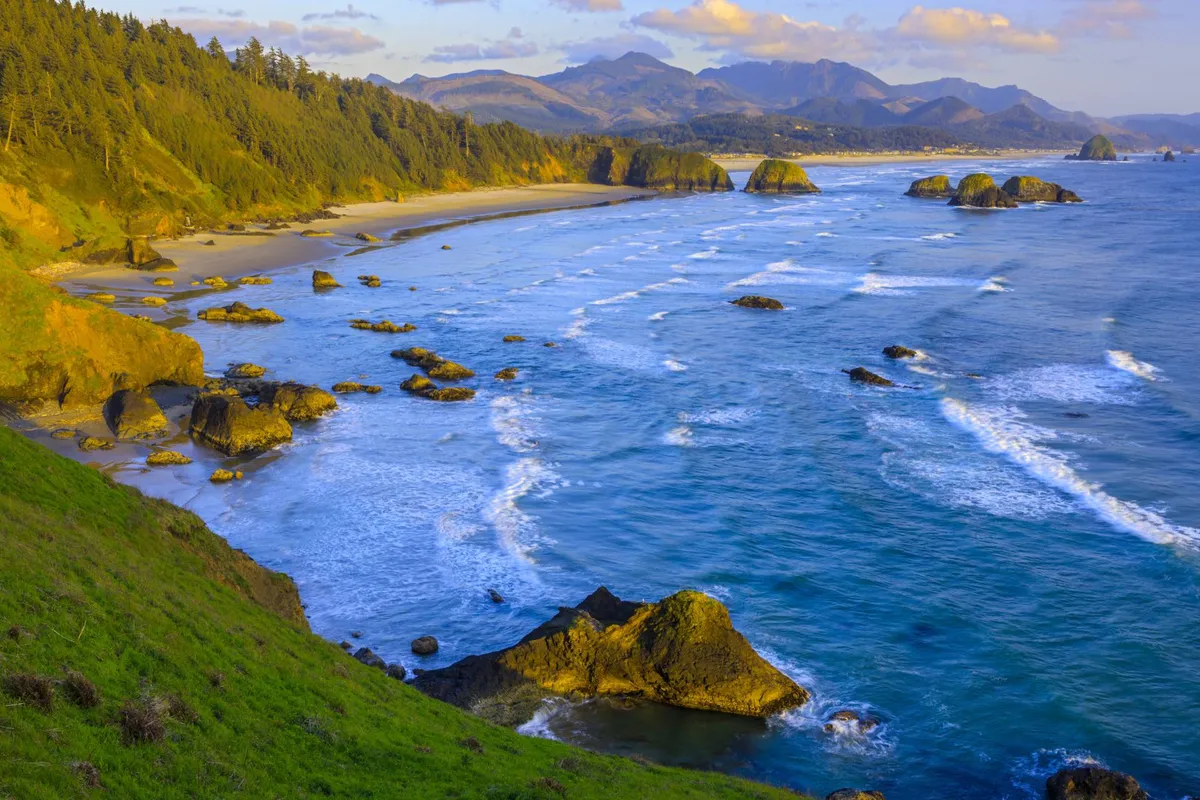 A sweeping coastal scene showcasing gentle ocean waves breaking onto a sandy beach, framed by striking rock formations and distant mountains under an open sky. The pristine natural beauty invites contemplation, relaxation, and outdoor adventure.